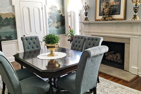 A square dark wood dining table with a potted fern centerpiece sits in a historic antebellum inn in Columbia, TN, surrounded by four tufted chairs in a room with a fireplace and mural wallpaper.
