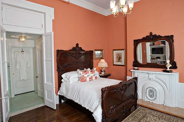 A coral-walled bedroom at a bed and breakfast in Columbia, TN, featuring a dark wood ornate bed frame, a white fireplace mantel with a decorative mirror, and a doorway to a dressing area with a white robe hanging on the door.