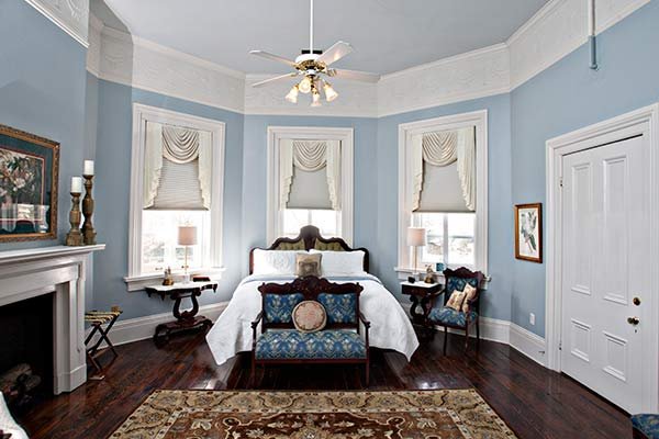 A bedroom with pale blue walls, a white bed, and a blue patterned settee, featuring three windows with draped valances, a fireplace, and a ceiling fan in one of our Columbia, TN, bed and breakfast guest rooms.