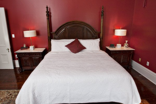 A bed with a dark wood headboard, white bedding, and a single maroon accent pillow, centered between two nightstands with lit lamps against a deep red wall in this Columbia, TN bed and breakfast guest room.