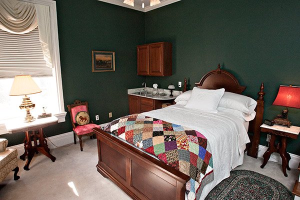 A cozy bedroom with dark green walls, a wooden bed featuring a patchwork quilt, a small sink area, and two side tables with lamps in a Columbia, TN bed and breakfast.