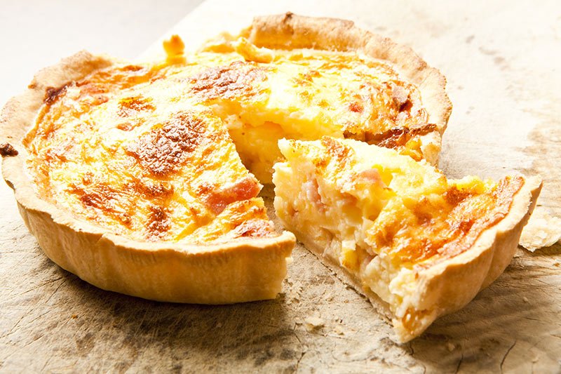 A golden-brown gourmet breakfast quiche with one slice cut and slightly separated on a wooden board.
