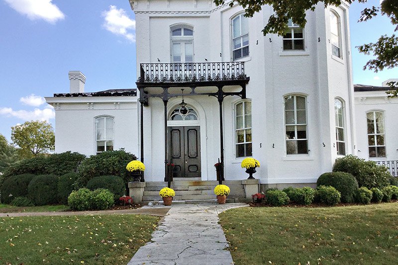 A white brick historic antebellum inn in Columbia, TN, featuring a dark front door, a wrought-iron balcony, and yellow mums in planters along the front steps.