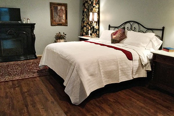 A guest room in Columbia, TN, featuring a queen bed with a white quilt and a maroon runner, positioned near a black fireplace and a wood-patterned floor.
