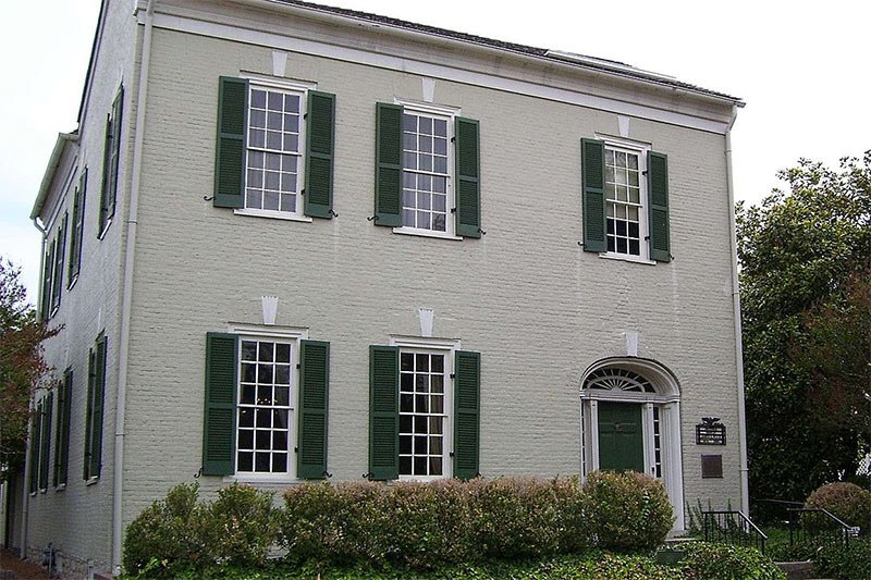 A two-story, light-colored brick house in Columbia, TN, with green shutters, a dark front door, and a historic plaque near the entrance, often passed by those attending local events.
