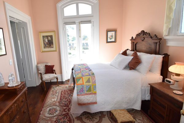 A bedroom with a wooden bed frame, a patchwork quilt, and light pink walls, featuring a large windowed door and a patterned area rug in a Columbia, TN bed and breakfast.