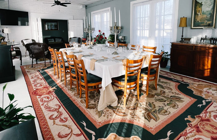 A long dining table set for twelve with a white tablecloth, tall candles, and floral centerpieces, surrounded by wooden chairs on a large patterned rug at a Columbia, TN bed and breakfast.